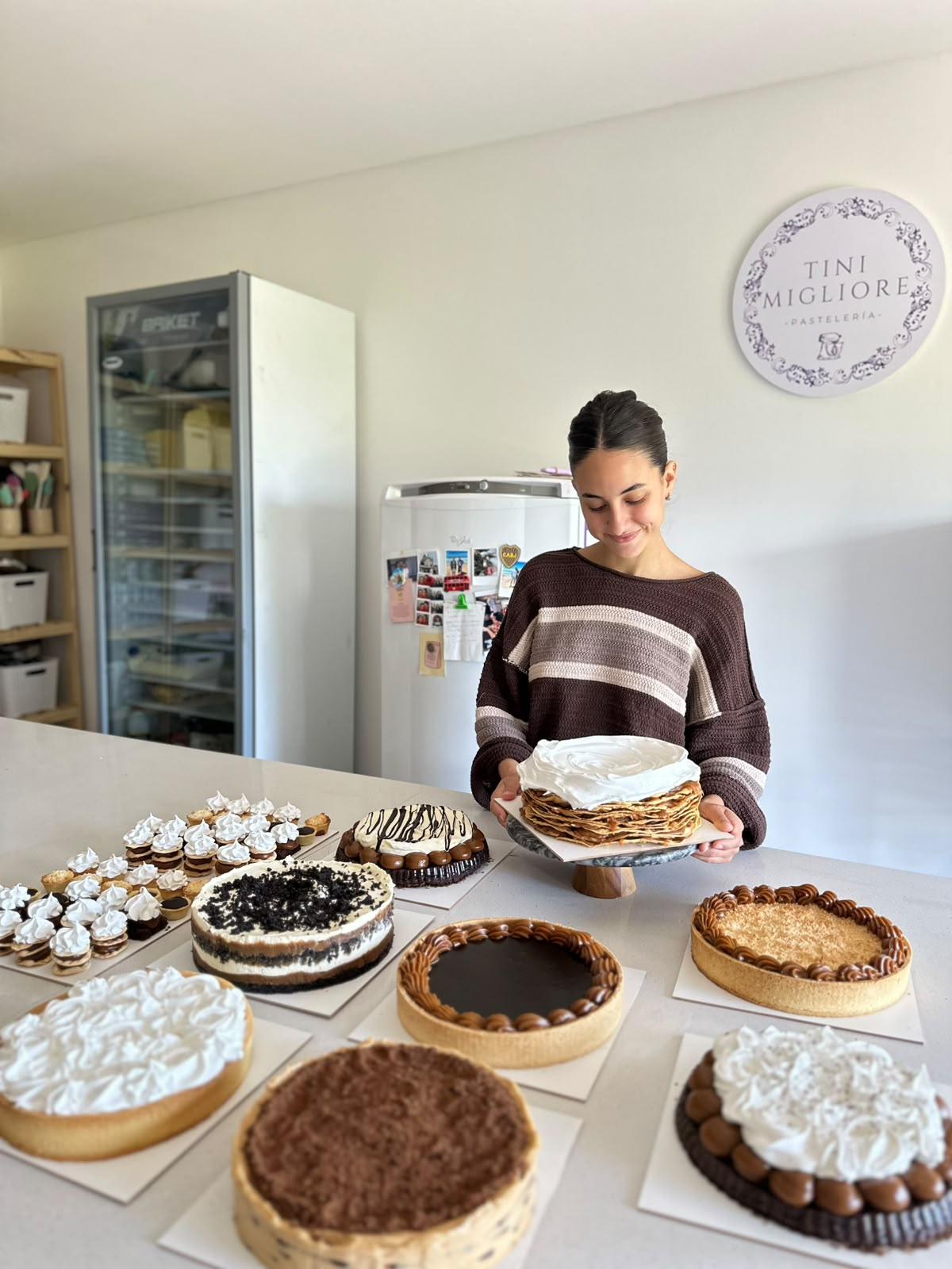Tini Migliore en su taller, sosteniendo una torta rodeada de su producción de pastelería